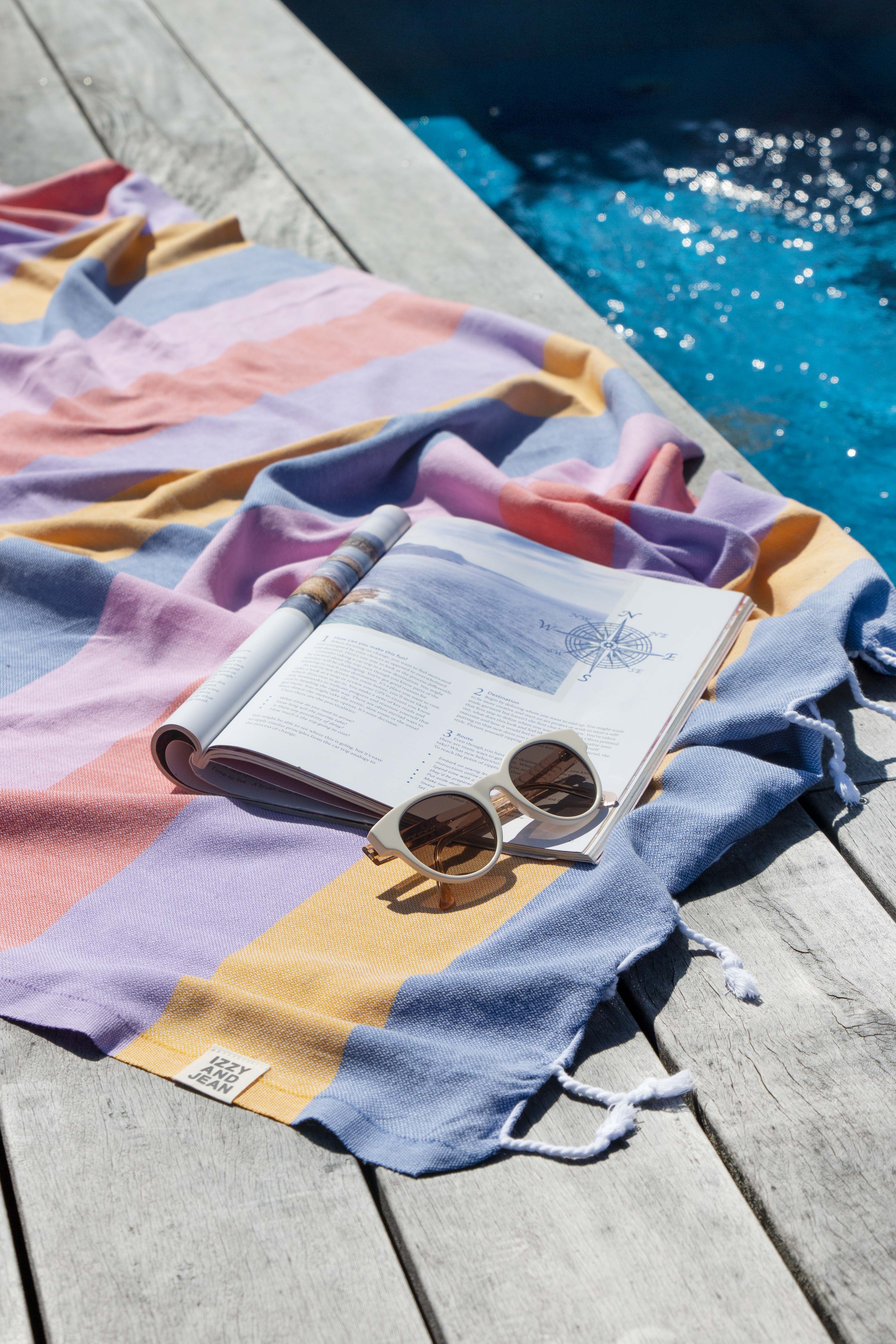A striped multi-coloured turkish towel in shades of blue, pink, and orange is on the side of a pool with an open magazine and sunnies on it