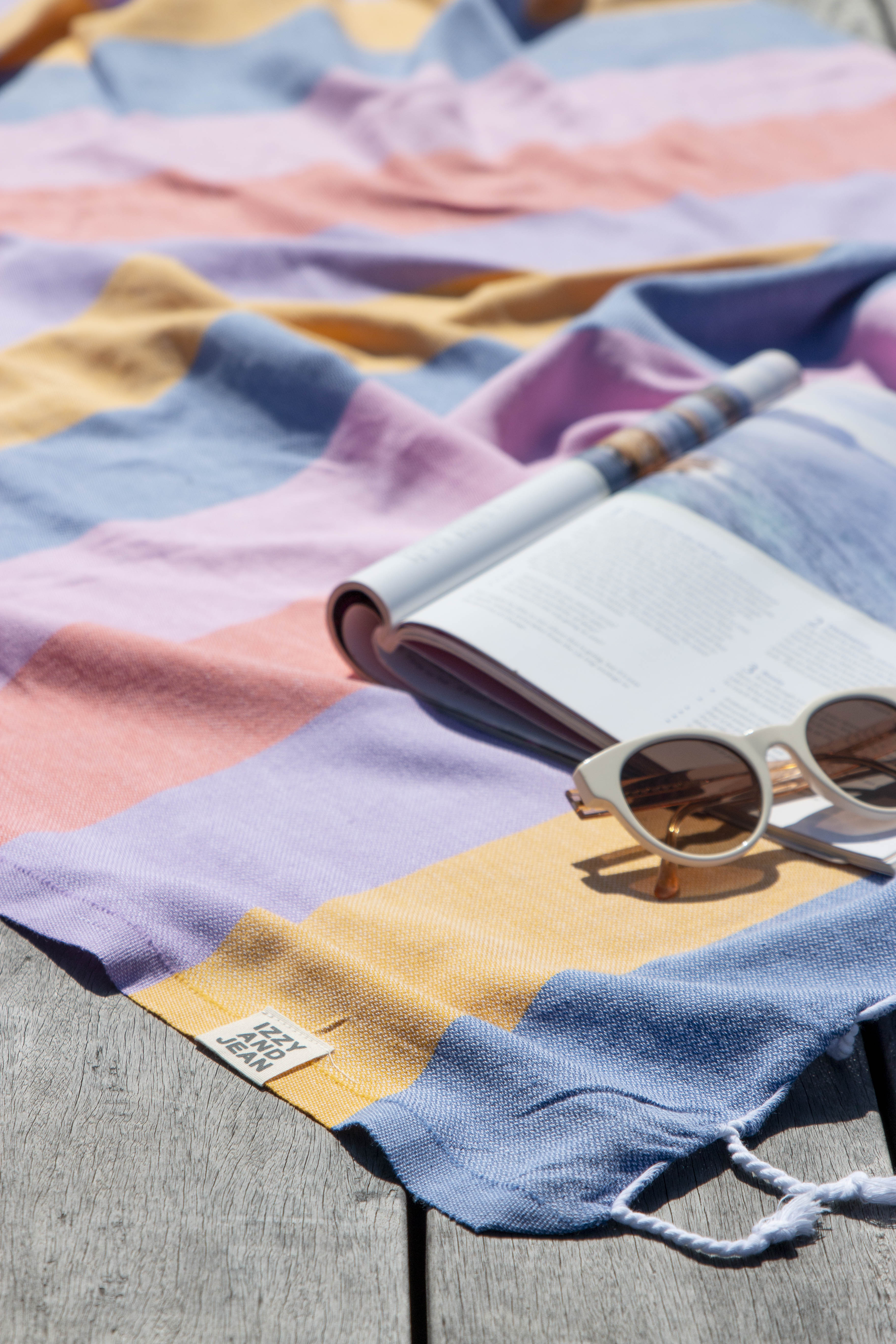 A striped multi-coloured turkish towel in shades of blue, pink, and orange on the ground with sunglasses and an open magazine indicating someone has been sunbathing.