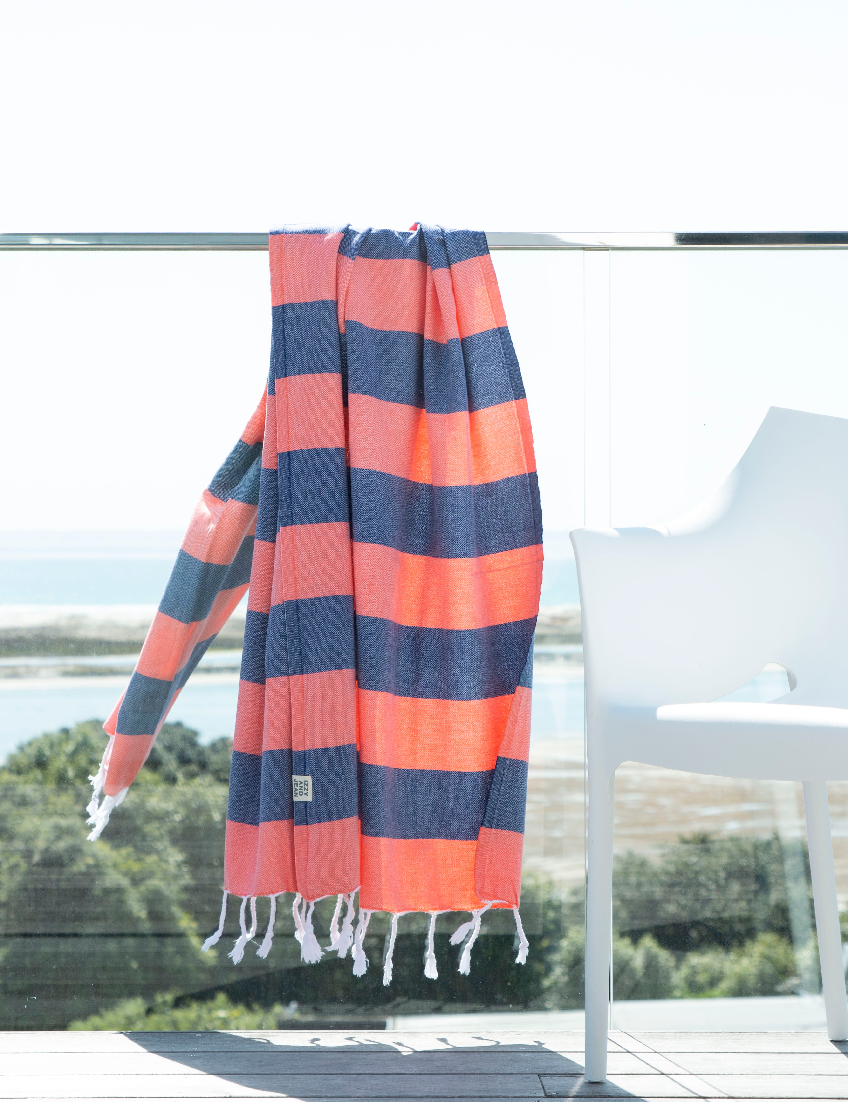 A Santorini Navy and Coral Striped Turkish Towel lying on a glass banister over-lookng a beach