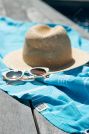 A marine (blue) coloured Classic Turkish towel with white stripes and fringes lays beside a pool on a wooden deck, with a straw hat and white sunglasses on top of it