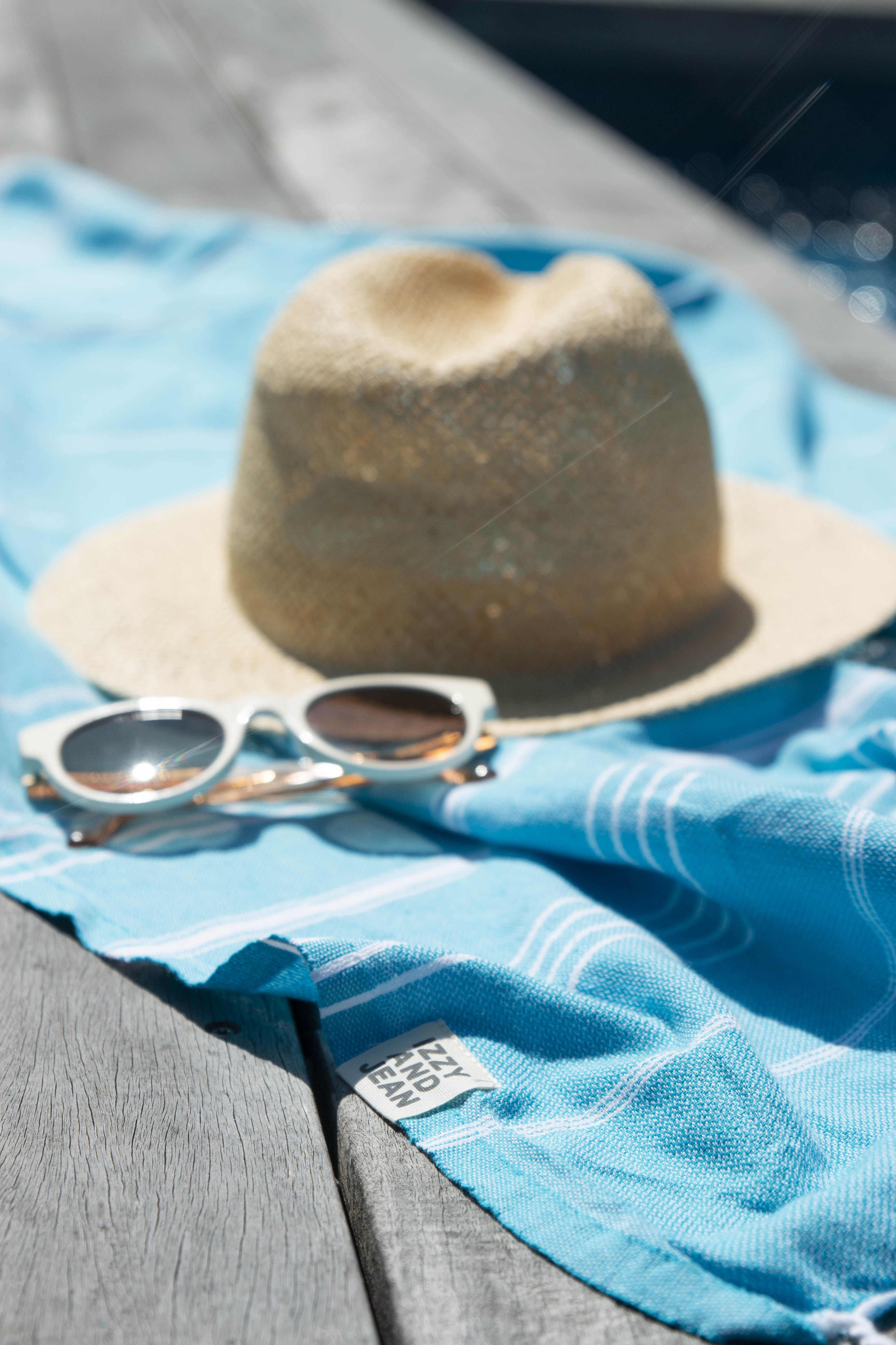 A marine (blue) coloured Classic Turkish towel with white stripes and fringes lays beside a pool on a wooden deck, with a straw hat and white sunglasses on top of it