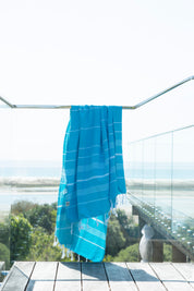 A marine-coloured Classic Turkish towel with white stripes and fringes hangs on a glass banister on a deck overlooking a beautiful ocean vista