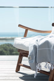 A pale grey Turkish towel with white stripes and tassels on the bottom edge lies over a stylish wooden chair, which is on a deck with a beautiful ocean vista in the background