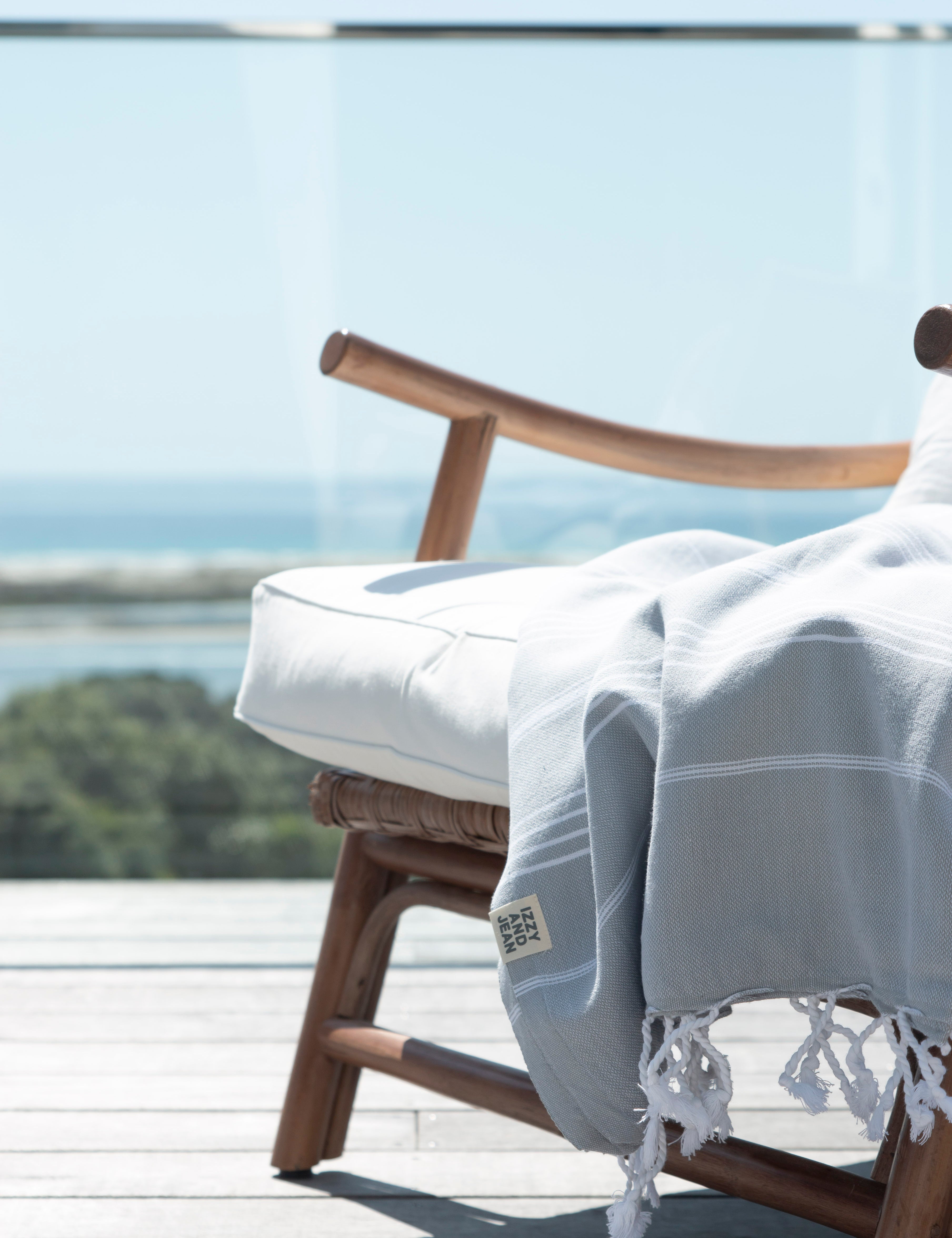 A pale grey Turkish towel with white stripes and tassels on the bottom edge lies over a stylish wooden chair, which is on a deck with a beautiful ocean vista in the background