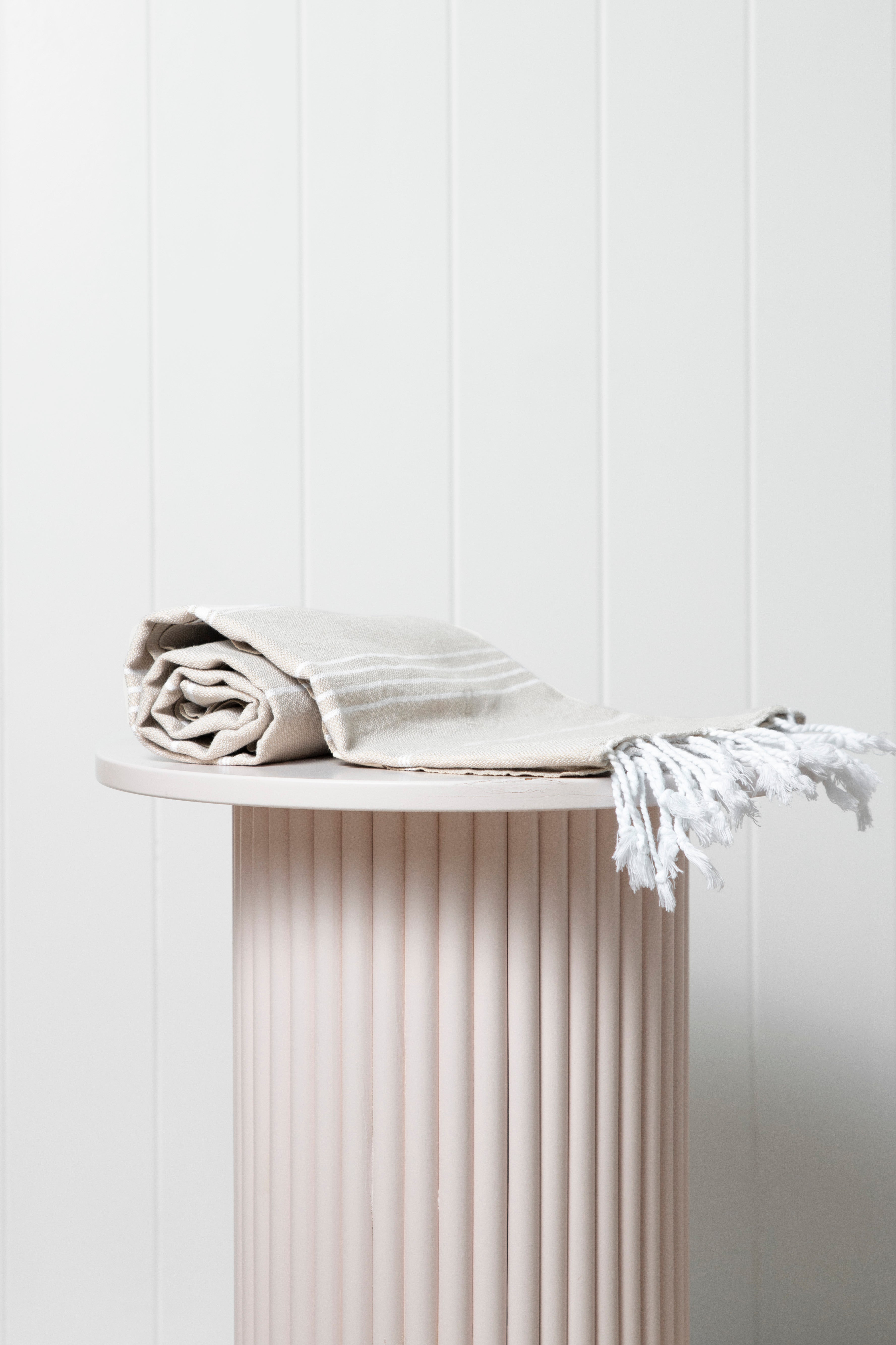 A sand Turkish towel with white stripes and tassels on the bottom edge on a pedestal with white wooden walls behind it