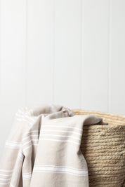 A sand Turkish towel with white stripes and tassels on the bottom edge draped over a weave bag