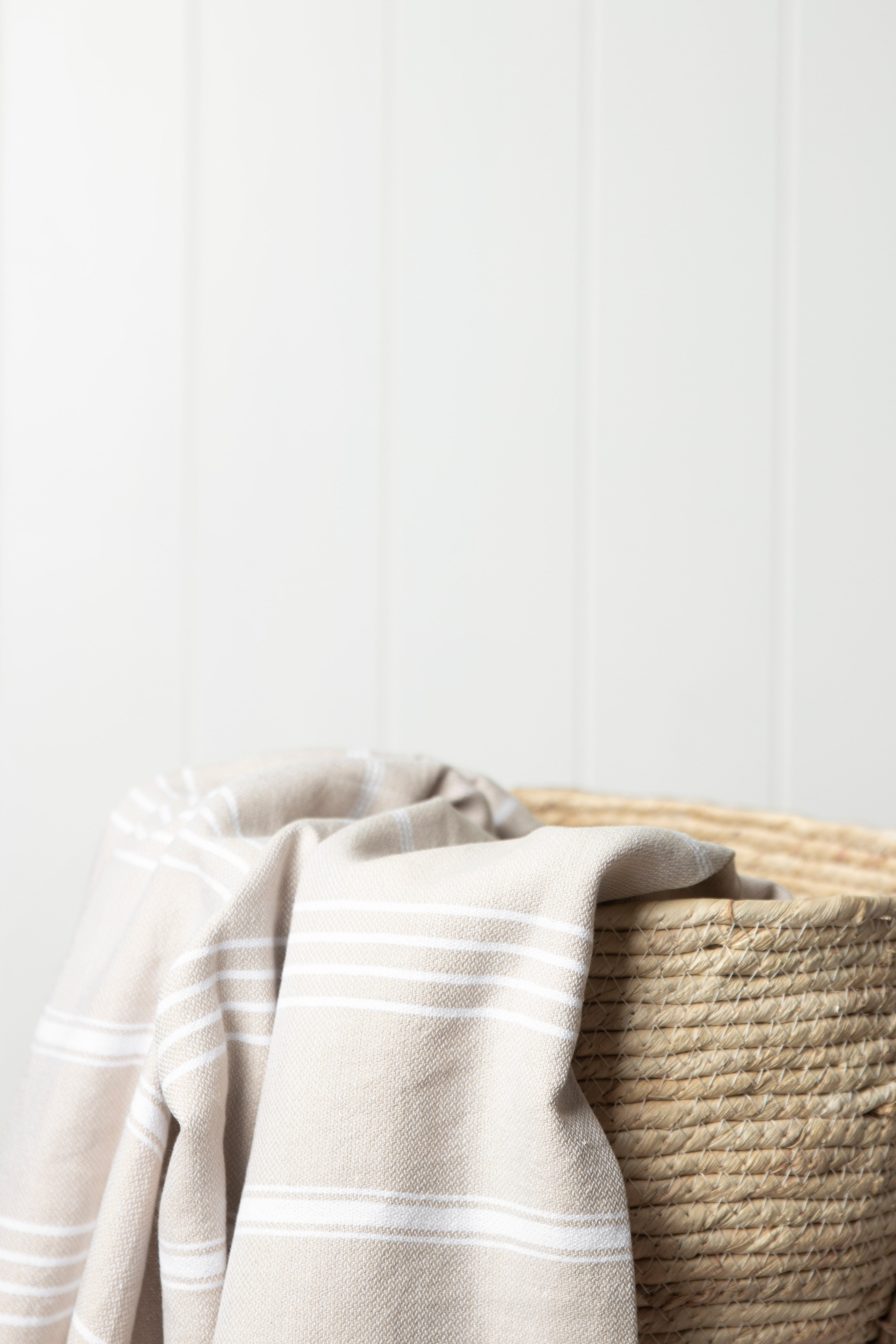 A sand Turkish towel with white stripes and tassels on the bottom edge draped over a weave bag