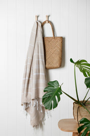 A sand Turkish towel with white stripes and tassels on the bottom edge hangs on a hook with white wooden walls and a weave bag behind it and a lush green plant on the ground near by.