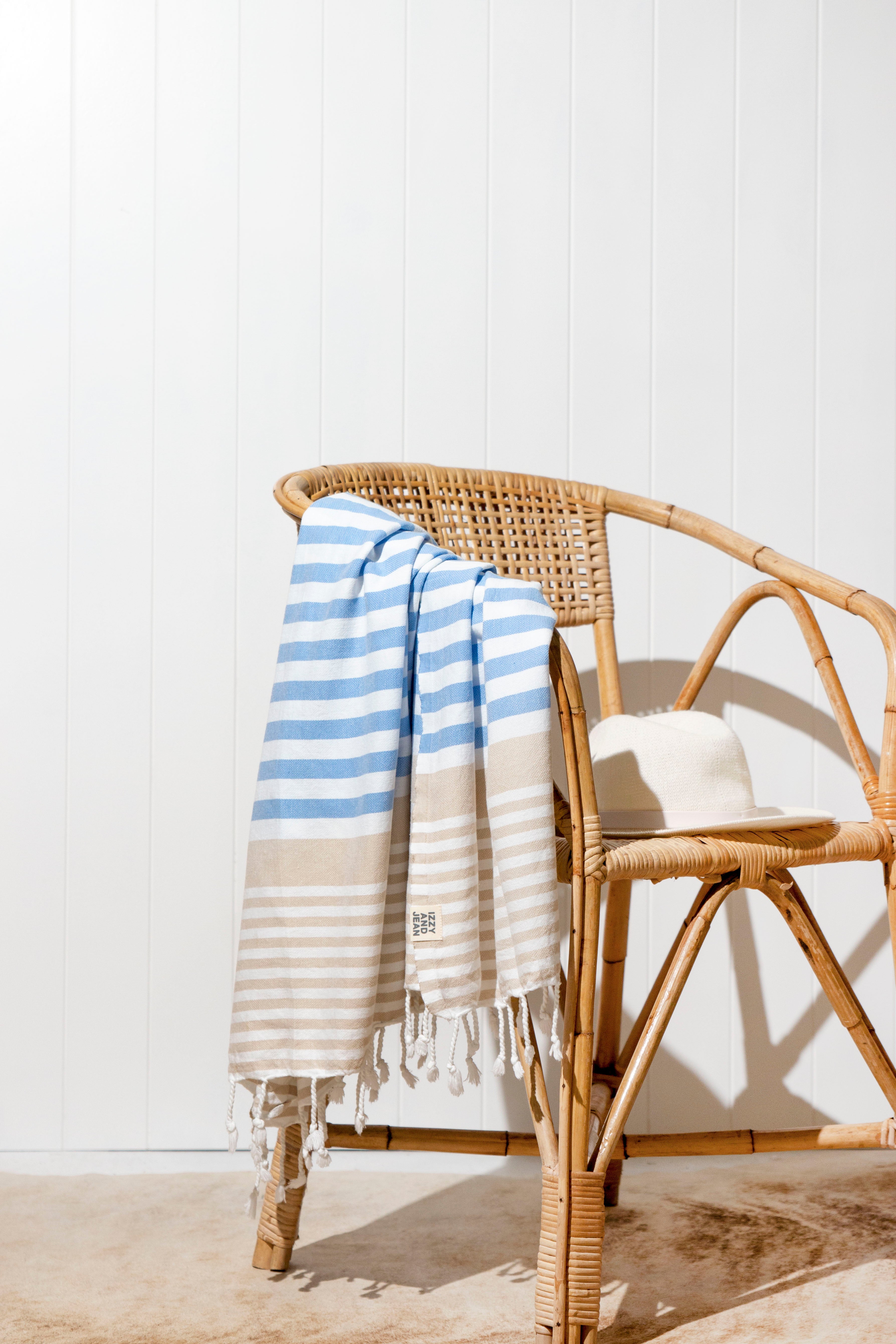 A draped Sofia Turkish towel with sky blue, white and sand stripes, and white fringes at the bottom hangs over a weaved outdoor chair with a sunhat next to it