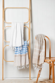 The Sofia Turkish towel in sky blue, white and sand stripes, with white fringes at the bottom hangs off a wooden ladder
