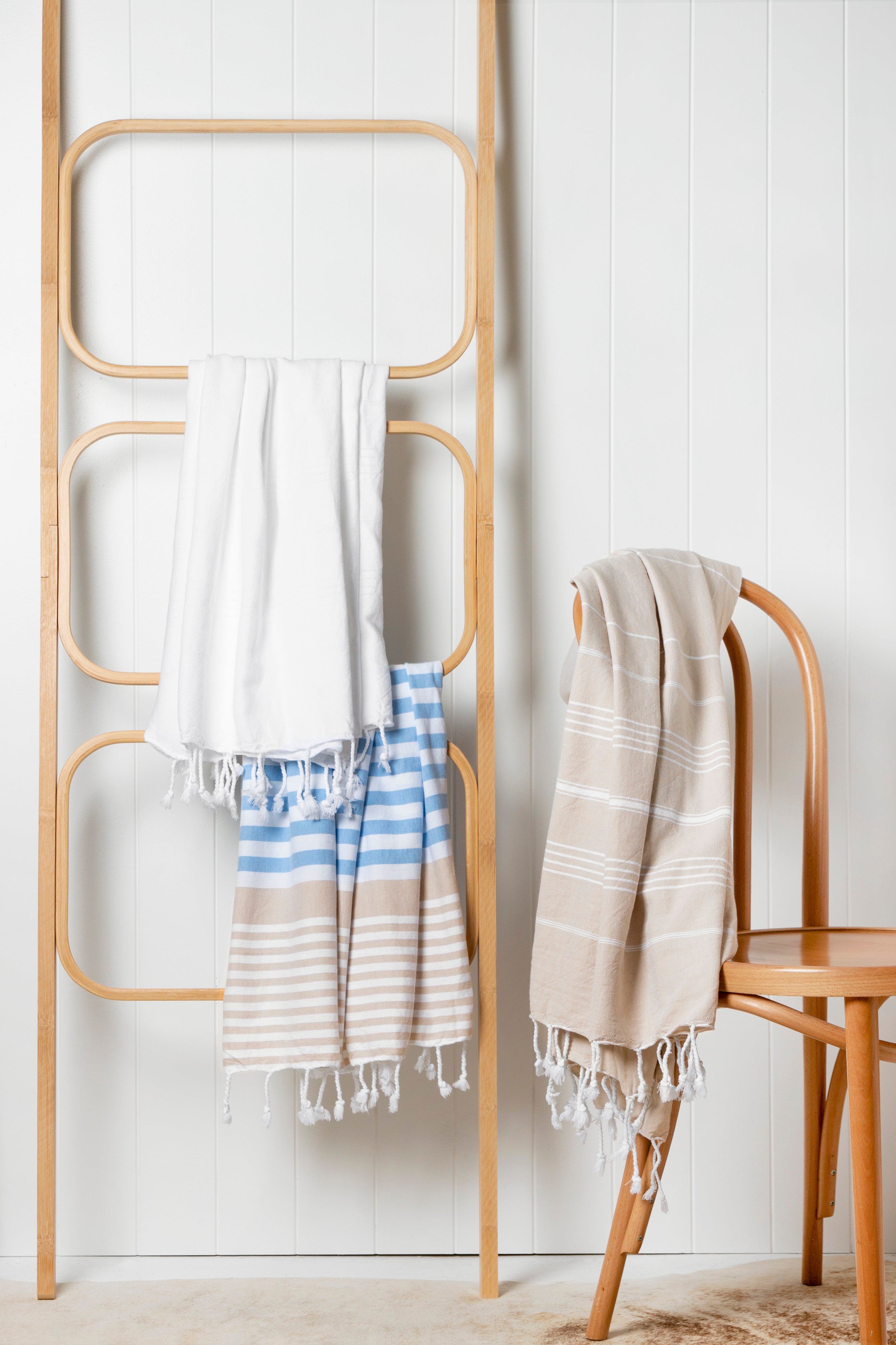 The Sofia Turkish towel in sky blue, white and sand stripes, with white fringes at the bottom hangs off a wooden ladder