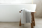 A pale grey coloured waffle towel lying on a wooden stool in front of a large white luxury bath in a bathroom