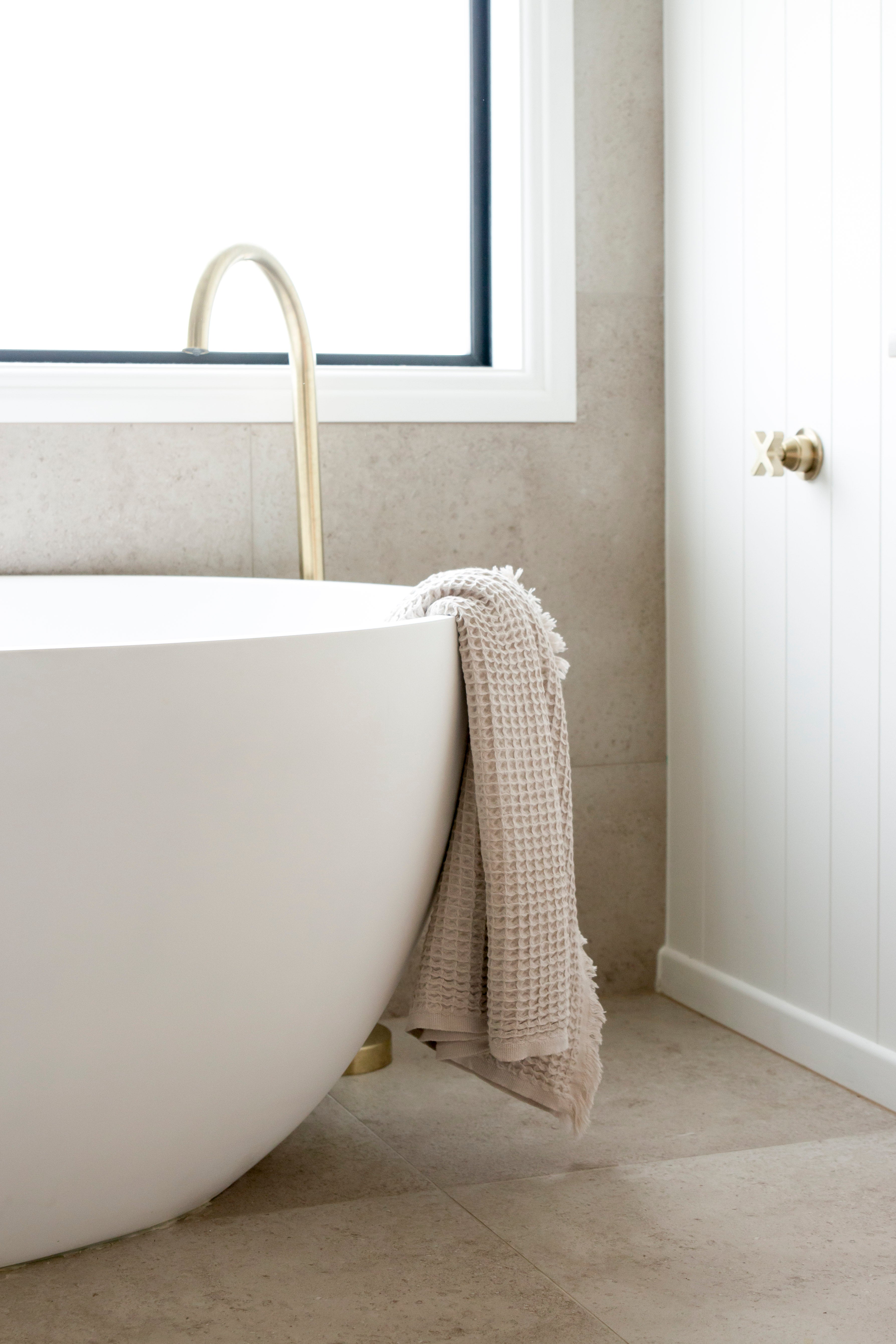 An oatmeal coloured waffle bath towel with tassles on the end, lying over the end of a bath in a luxury bathroom.