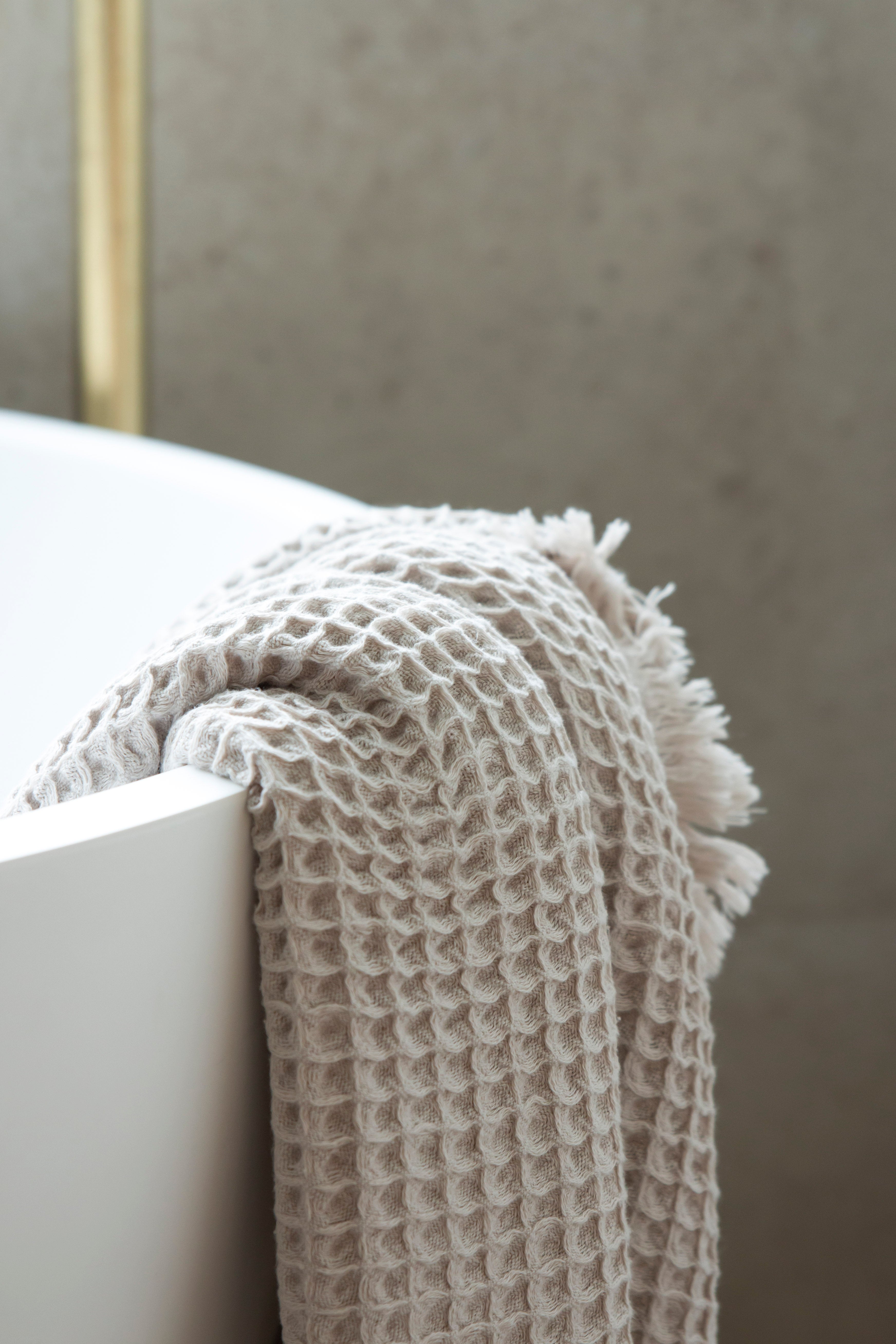 An oatmeal-coloured waffle bath towel with tassles, lying over the edge of a bath with a gold plated tap in the background.