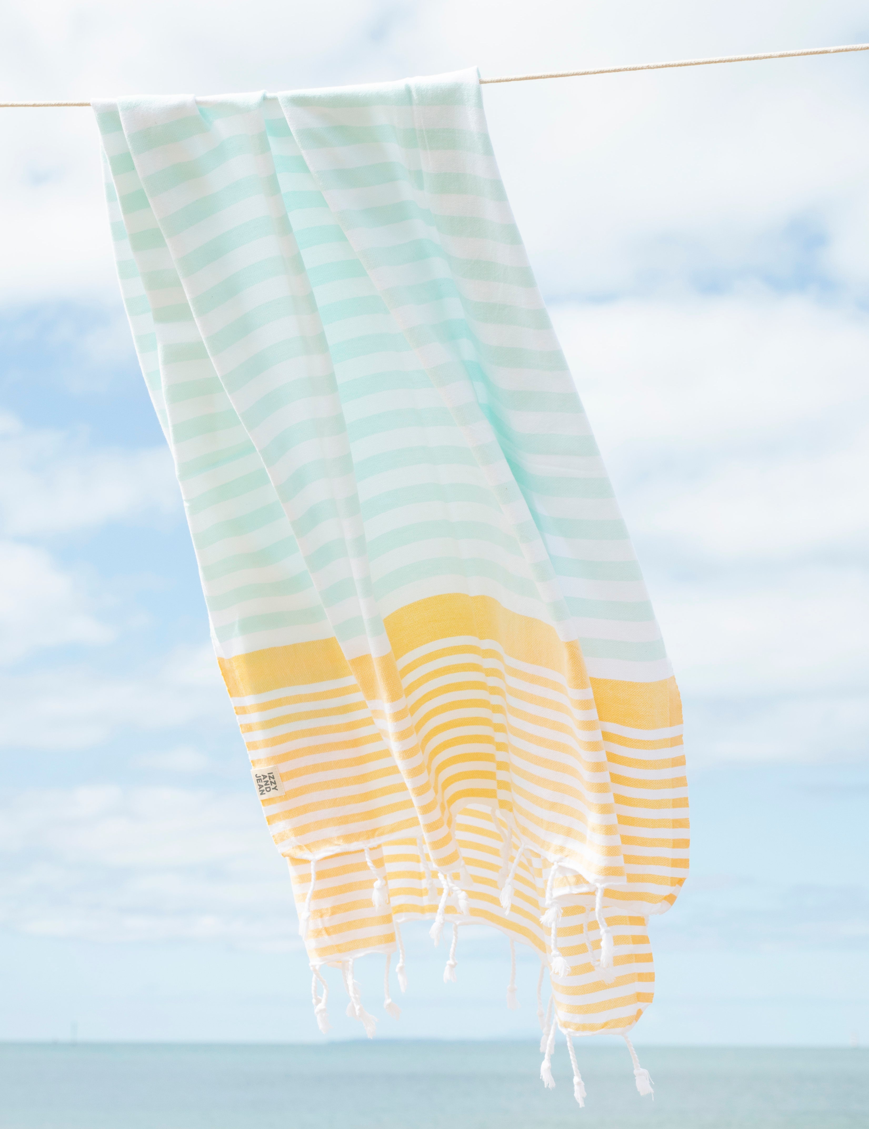 A mint, yellow and white striped Turkish beach towel hanging in the wind on a washing line with the beach in the background
