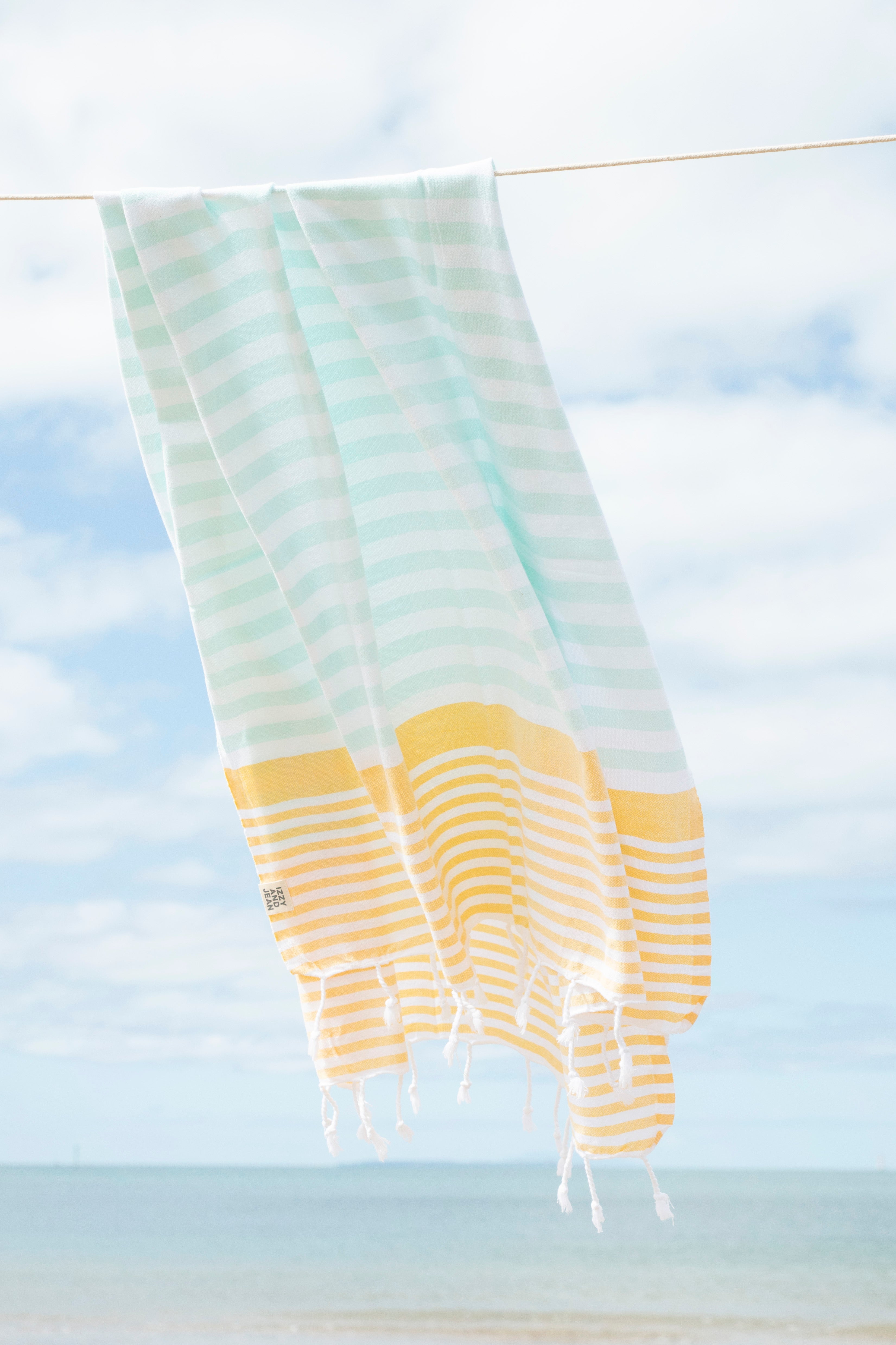 A mint, yellow and white striped Turkish beach towel hanging in the wind on a washing line with the beach in the background