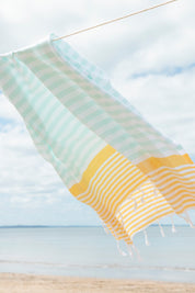 A mint, yellow and white striped Turkish towel blowing in the wind on a washing line with the beach and sand in the background