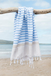 A striped Sofia Turkish towel in sky blue and sand, with white stripes and fringes at the bottom hangs from a tree branch with the beach in the background
