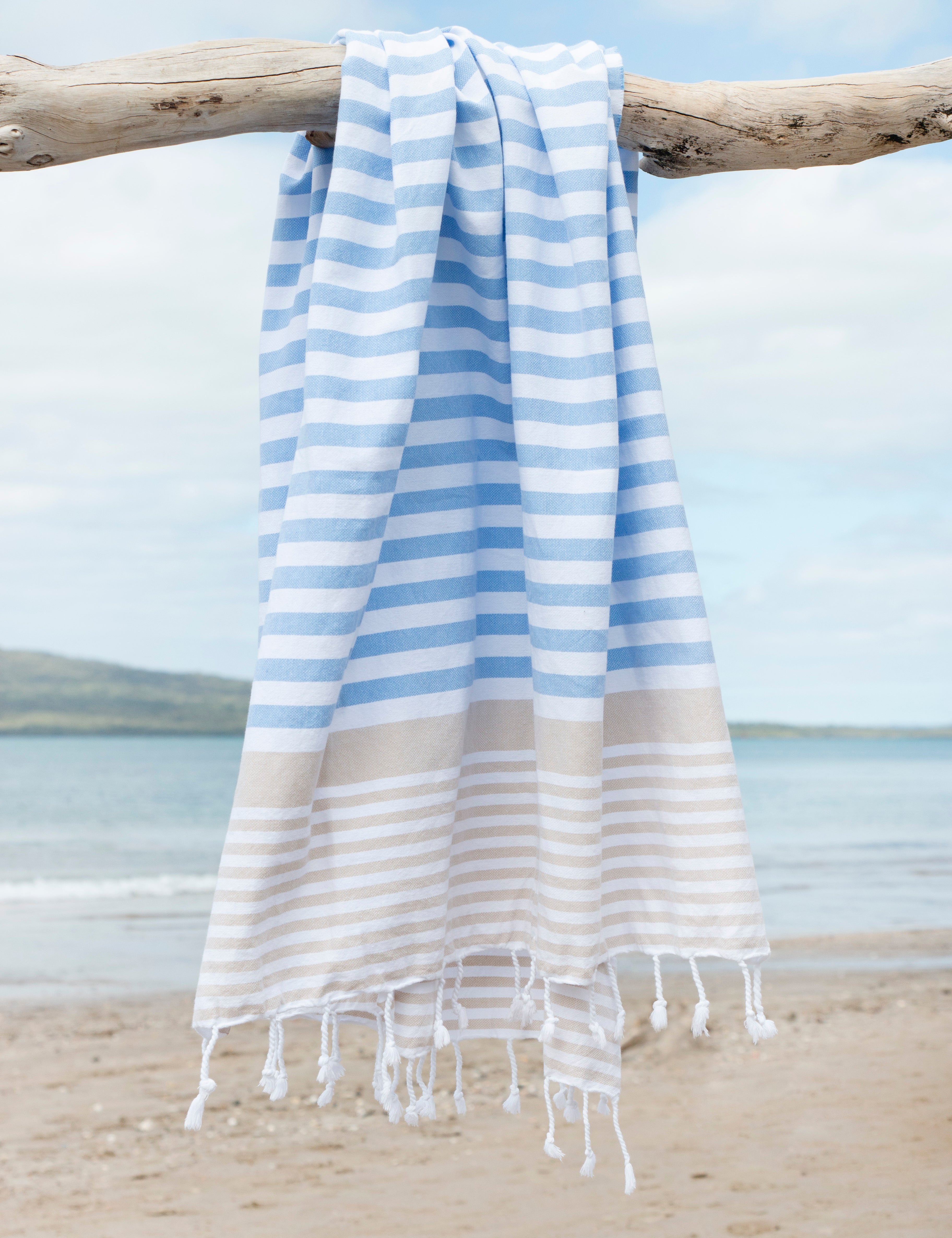 A striped Sofia Turkish towel in sky blue and sand, with white stripes and fringes at the bottom hangs from a tree branch with the beach in the background