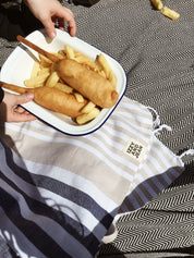Fish and Chipes on a plate sitting on a  Navy and Sand striped Sofia Turkish Towel with tassles at the bottom
