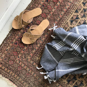 A black Turkish towel with white stripes and fringes on the bottom lying strewn on a Turkish rug with stylish sandals next to them