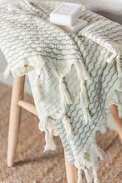 A silver-grey Anatolia waffle honeycomb weave with a white back and white tassels on a wooden stool with luxury soap on top.
