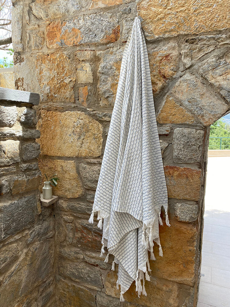 A silver-grey Anatolia waffle honeycomb weave with a white back and white tassels handing on a hook on a brick background