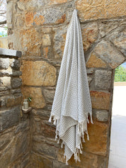 A silver-grey Anatolia waffle honeycomb weave with a white back and white tassels handing on a hook on a brick background