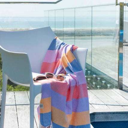 A striped multi-coloured turkish towel in shades of blue, pink, and orange is draped over a white chair, with a pool and balcony in the background.