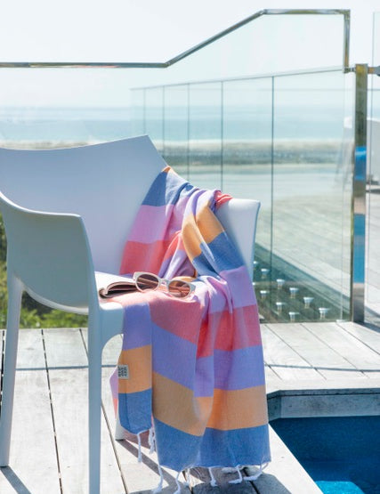 A striped multi-coloured turkish towel in shades of blue, pink, and orange is draped over a white chair, with a pool and balcony in the background.