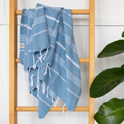 An Aegean Blue Turkish Towel hanging on a wooden ladder rung with a plant next to it
