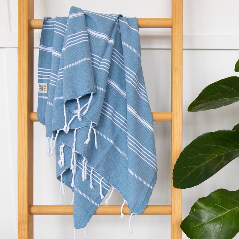 An Aegean Blue Turkish Towel hanging on a wooden ladder rung with a plant next to it