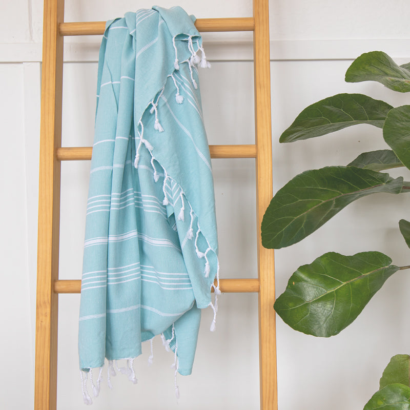 A mint Turkish towel with white stripes and fringes displayed hanging on a wooden ladder with a lush green plant beside it.