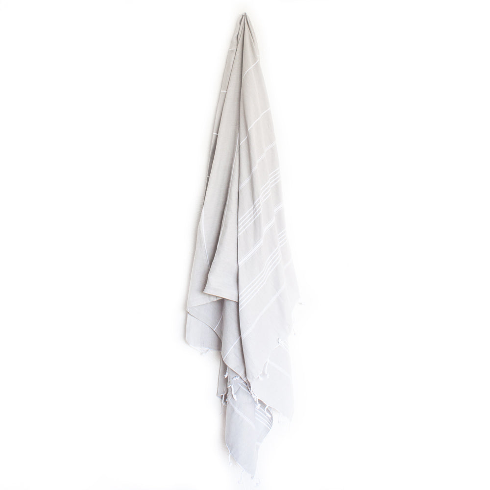 A pale grey Turkish towel with white stripes and tassels on the bottom edge hangs from a hook on a white background