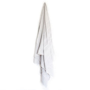 A pale grey Turkish towel with white stripes and tassels on the bottom edge hangs from a hook on a white background