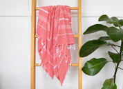 A watermelon Turkish towel with white stripes, displayed vertically with the fringes at the bottom hangs off a rung on a wooden ladder with a plant nearby