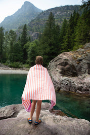 A boy wrapped in a coral and fuchsia striped Turkish beach towel on a rock looking into the lake water with pine trees and a mountain in the distance