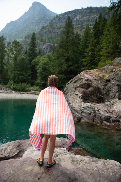 A boy wrapped in a coral and fuchsia striped Turkish beach towel on a rock looking into the lake water with pine trees and a mountain in the distance