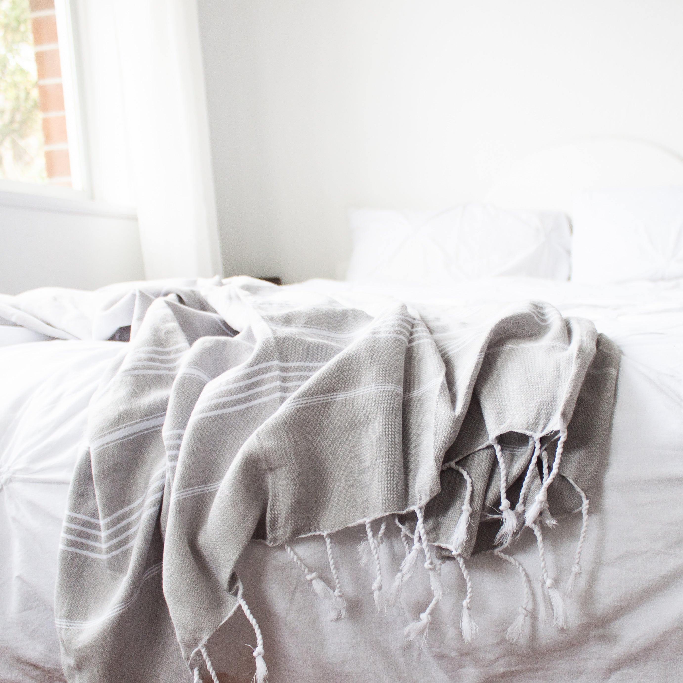 A pale grey Turkish towel with white stripes and tassels on the bottom edge, lies across an unmade bed with a white duvet