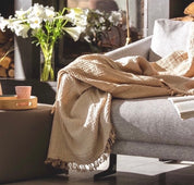 A mustard beige coloured 100% cotton woven blanket draped over the arm of a gray sofa, with a textured design and fringed edges.
