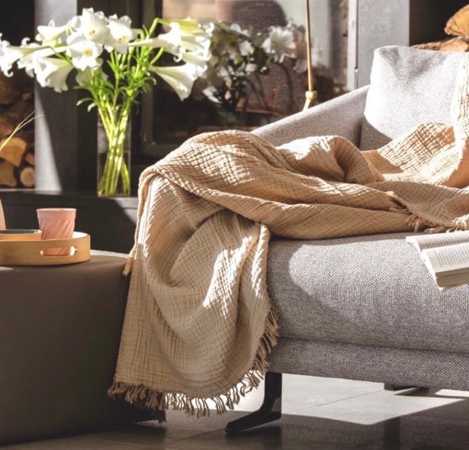 A mustard beige coloured 100% cotton woven blanket draped over the arm of a gray sofa, with a textured design and fringed edges.