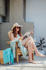 A lady with a straw hat sitting on a wooden chair reading a magazine with turquoise Turkish towel with white stripes, displayed drapped over a weave basket next to her.
