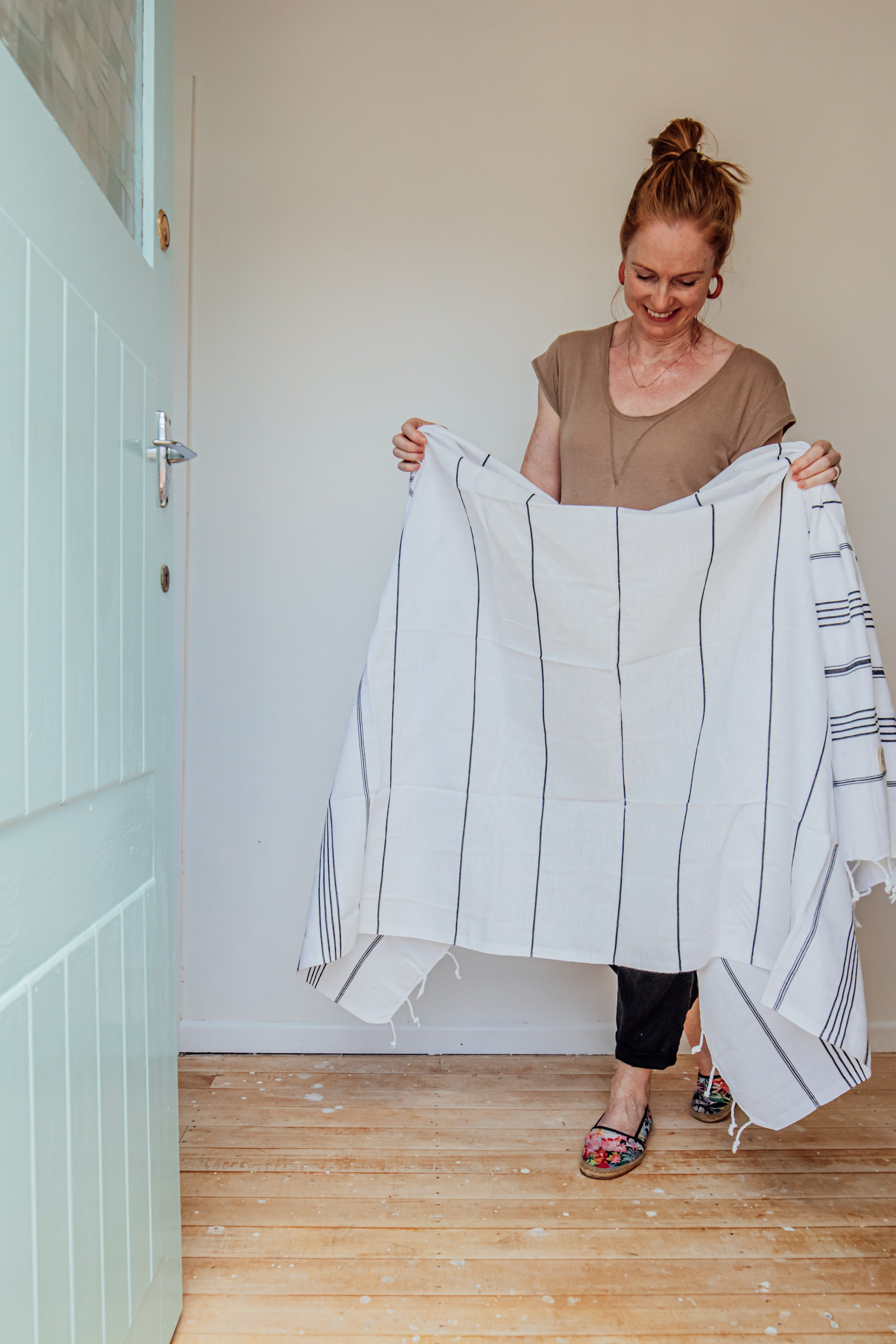 A smiling lady holds up a white Turkish towel with black stripes and tassels on the bottom edge inside her white wooden front door with wooden floors