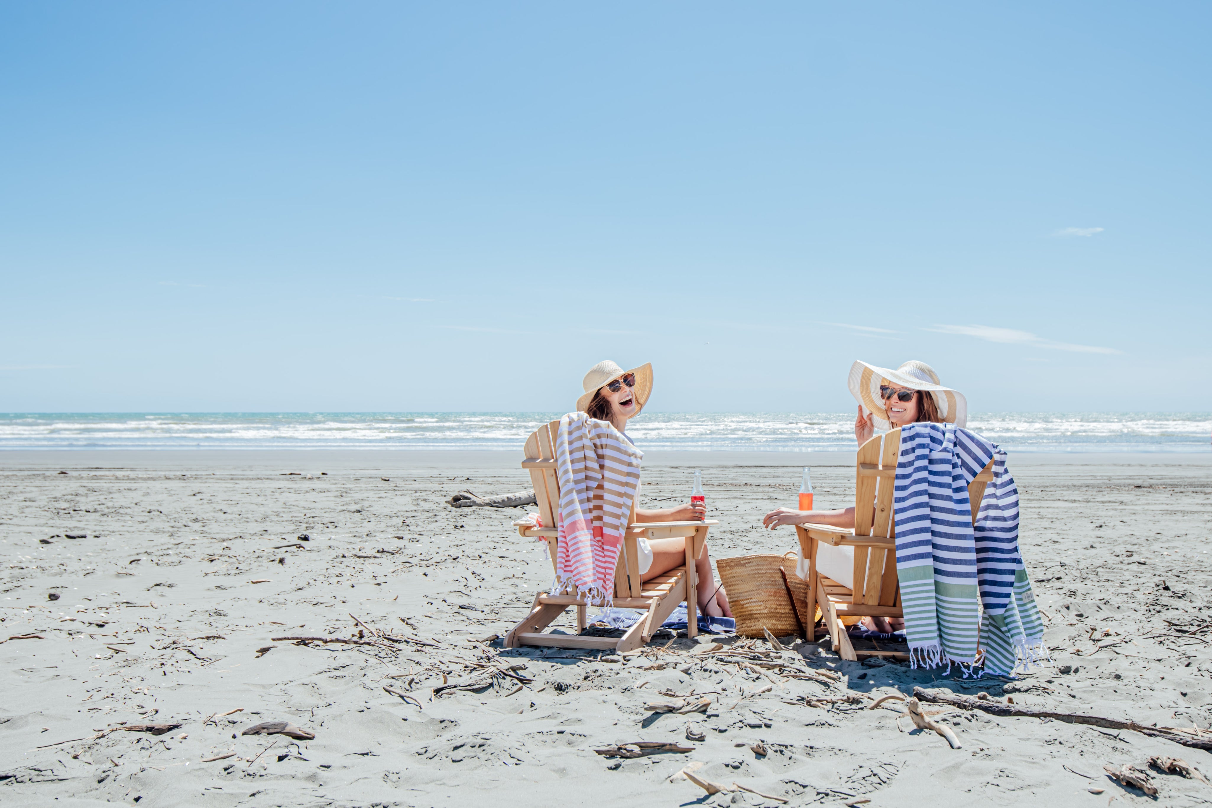 Ladies sitting on blue and green stripey Turkish towels on wooden beach chairs on the beach drinking champange