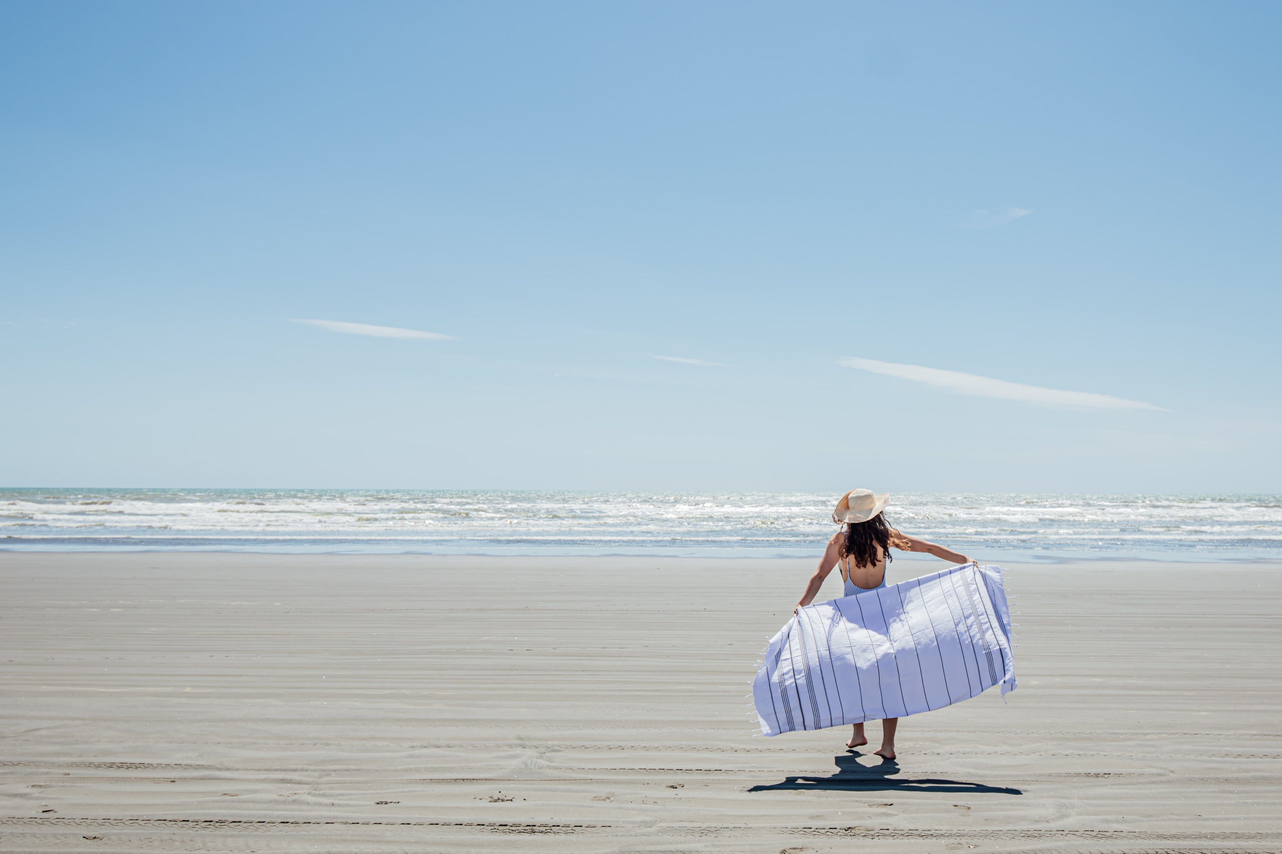 A beautiful beach scene with blue sky and a girl in a hat walks towards the sea away from the camera with a white Turkish towel bellowing out behind her