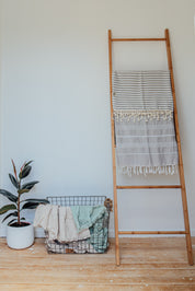 A pale grey Turkish towel with white stripes and tassels on the bottom edge folder over a rung on a wooden ladder
