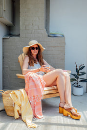  A lady sits on a wooden chair with a stylish straw hat and sunglasses, with a yellow Turkish towel draped over a weave basket next to her