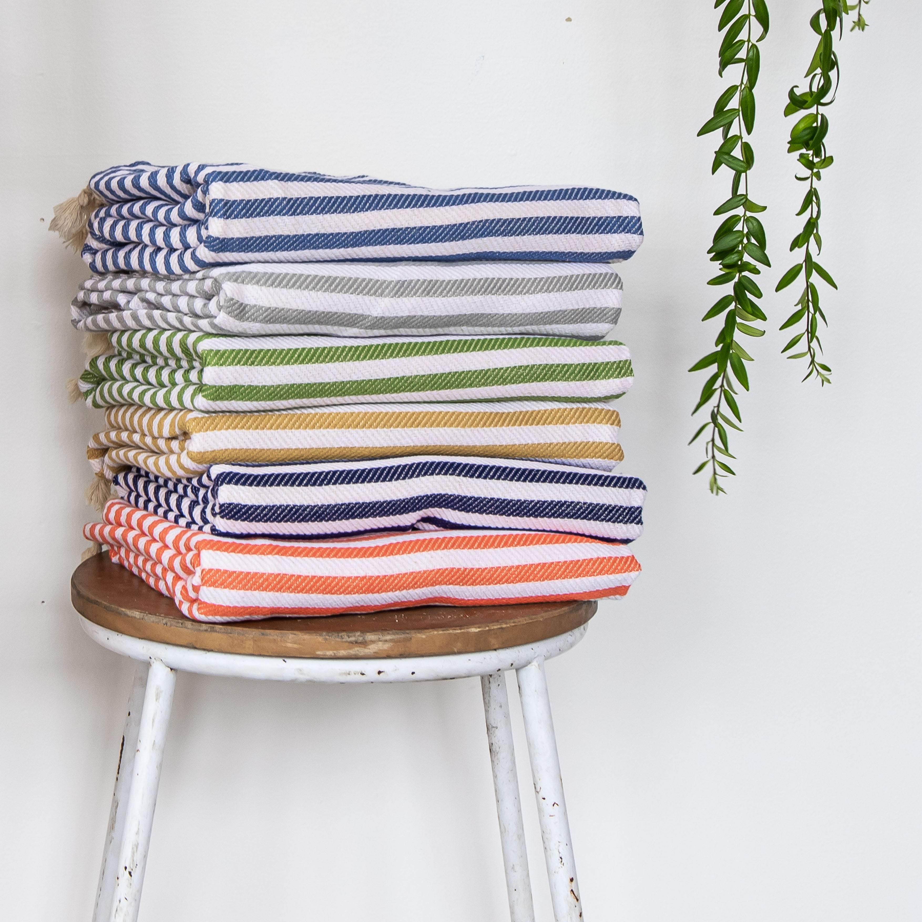 A range of coloured St Tropez turkish towels stacked up on top of a stool, in navy, grey, ocean, mustard, denim and orange.
