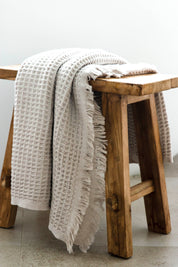 An oatmeal coloured waffle bath towel with tassles, lying over a wooden stool.
