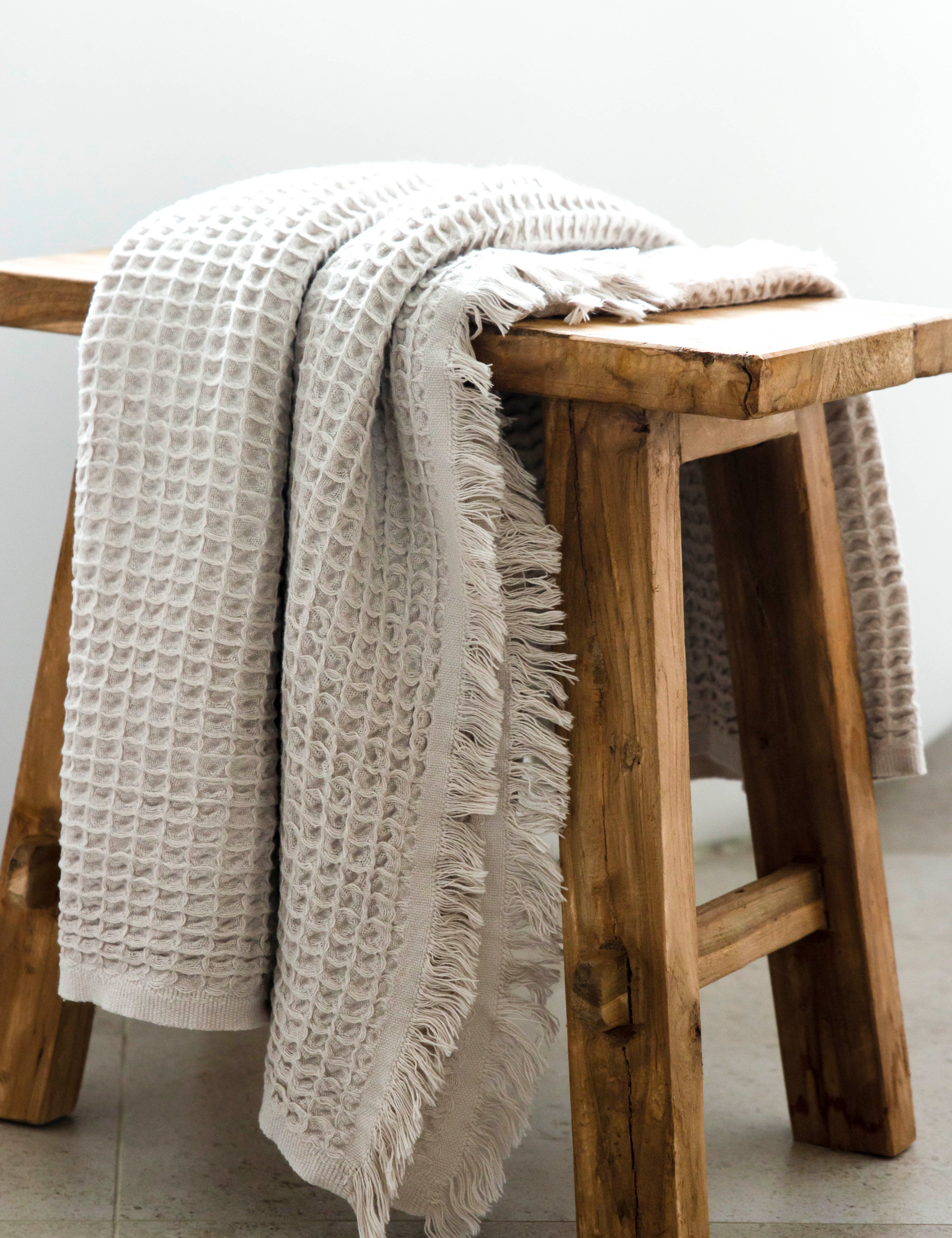 An oatmeal coloured waffle bath towel with tassles, lying over a wooden stool.